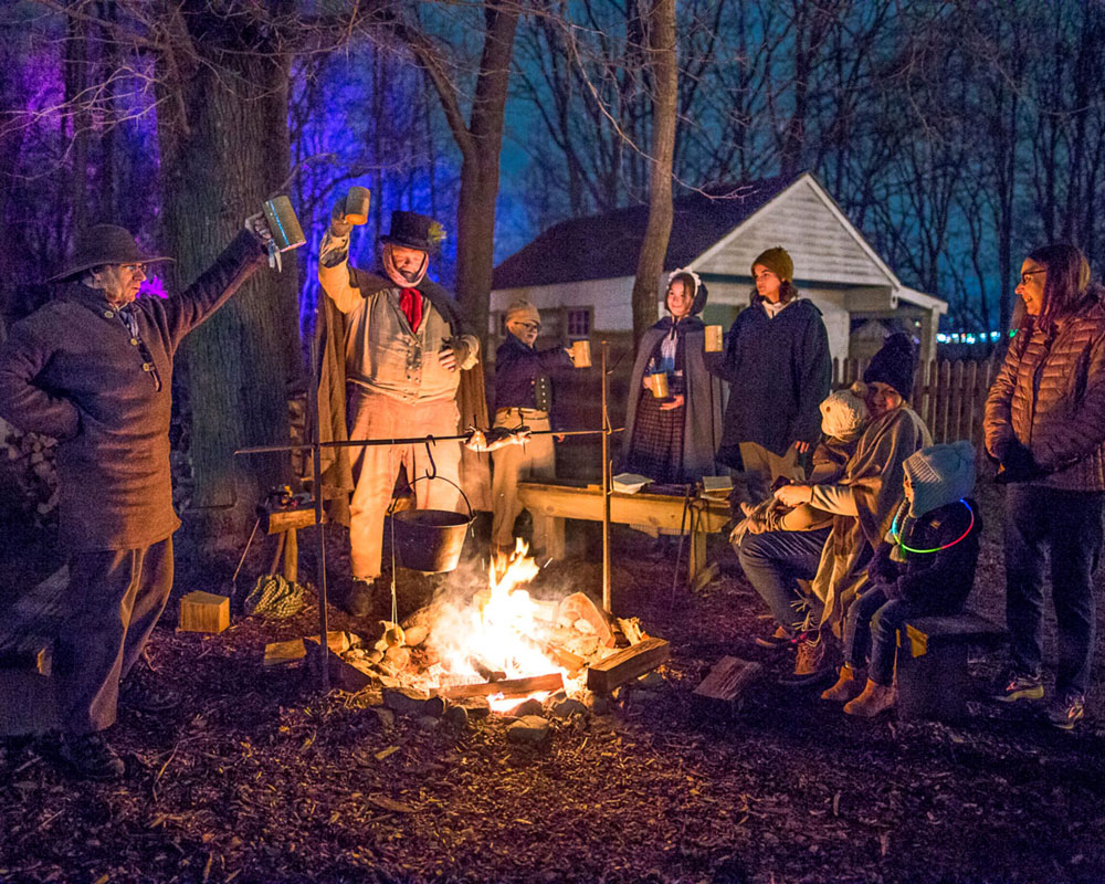 People gathered around a campfire. Two men are lifting up mugs in celebration.